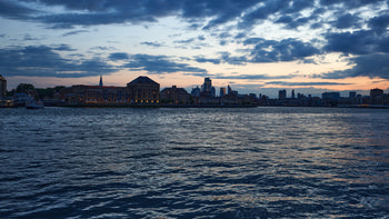 This urban landscape photograph captures Columbia Wharf at dusk during the summer evening in London. The image features the architecture of Columbia Wharf prominently along the banks of the River Thames, with the city skyline visible in the background, including notable landmarks such as The Shard. The gentle light from the setting sun reflects on the Thames, enhancing the atmosphere of this London riverside scene. The photograph highlights the contrast between the historic architecture of Columbia Wharf and the modern urban structures that define the London skyline.